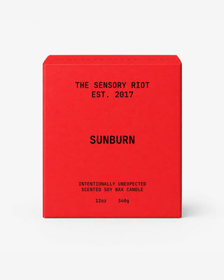 A red rectangular box for a scented soy wax candle with black text reading 'THE SENSORY RIOT EST. 2017', 'SUNBURN', and 'INTENTIONALLY UNEXPECTED SCENTED SOY WAX CANDLE 12oz 340g' on a light gray background.