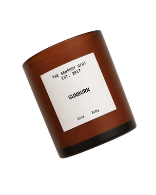 An amber glass jar candle with a white label reading 'THE SENSORY RIOT EST. SUNBURN' on a plain white background.