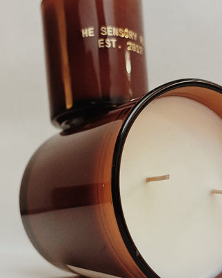 Amber glass candle jar with white wax and two wicks, shown with part of the matching amber container visible behind it on a light neutral background.