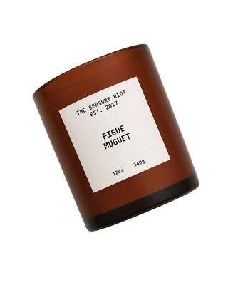 An amber glass jar candle with a white label reading 'THE SENSORY RIOT EST. 2017 FIGUE MUGUET' on a plain white background.