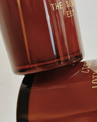 Close-up of an amber brown glass bottle with gold lettering, showing part of the label text 'THE SENIOR EST.' and 'LOTUS' on a light neutral background.