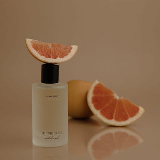 A frosted glass bottle of Hippie Soul Ritual Spray with a black cap, topped with a grapefruit slice, with whole and sliced grapefruit in the background against a warm beige backdrop.