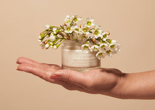 Hand holding a jar of cream with flowers on top against a beige background