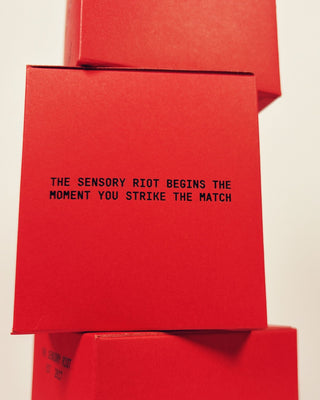 A close-up of a bright red square box with black printed text reading 'THE SENSORY RIOT BEGINS THE MOMENT YOU STRIKE THE MATCH' stacked on top of other matching red boxes against a plain light background.