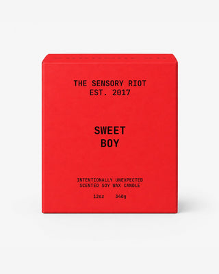 A bright red rectangular box for a scented soy wax candle, with black text reading 'THE SENSORY RIOT EST. 2017', 'SWEET BOY', and 'INTENTIONALLY UNEXPECTED SCENTED SOY WAX CANDLE 12oz 340g' on a light gray background.